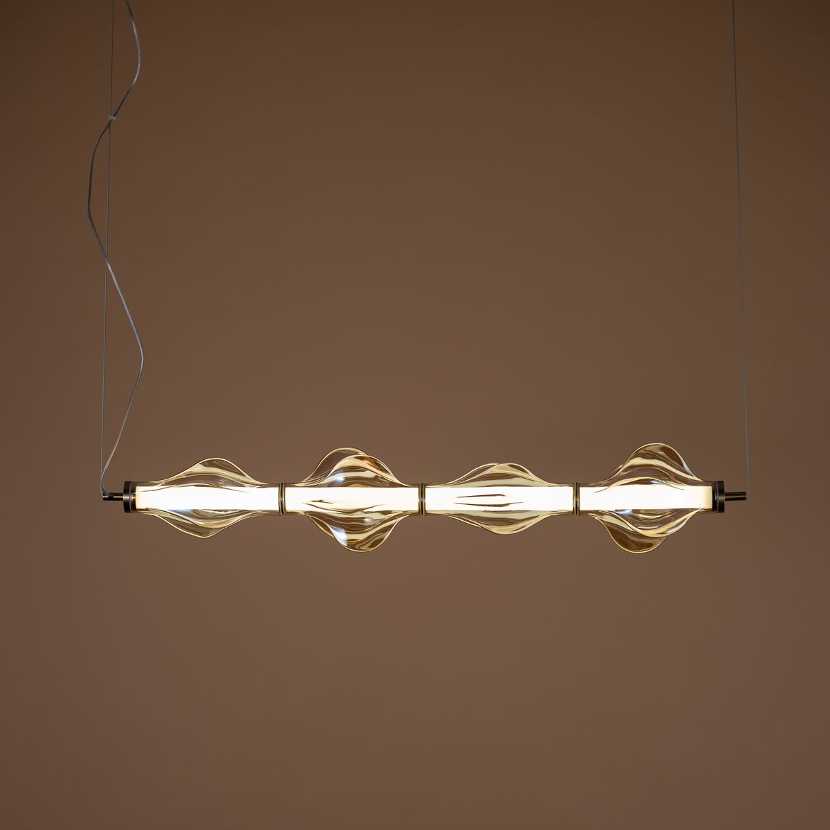 Lampa wisząca LED Elva 30W, kolor mosiężny z bursztynowymi detalami. Ma owalne klosze w kształcie fal, zamontowana w pozycji poziomej. Emituje ciepłe światło 3000K, zawieszona na cienkich linkach na tle jednolitej, ciepłej ściany.