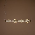 Lampa wisząca LED Elva 30W, kolor mosiężny z bursztynowymi detalami. Ma owalne klosze w kształcie fal, zamontowana w pozycji poziomej. Emituje ciepłe światło 3000K, zawieszona na cienkich linkach na tle jednolitej, ciepłej ściany.
