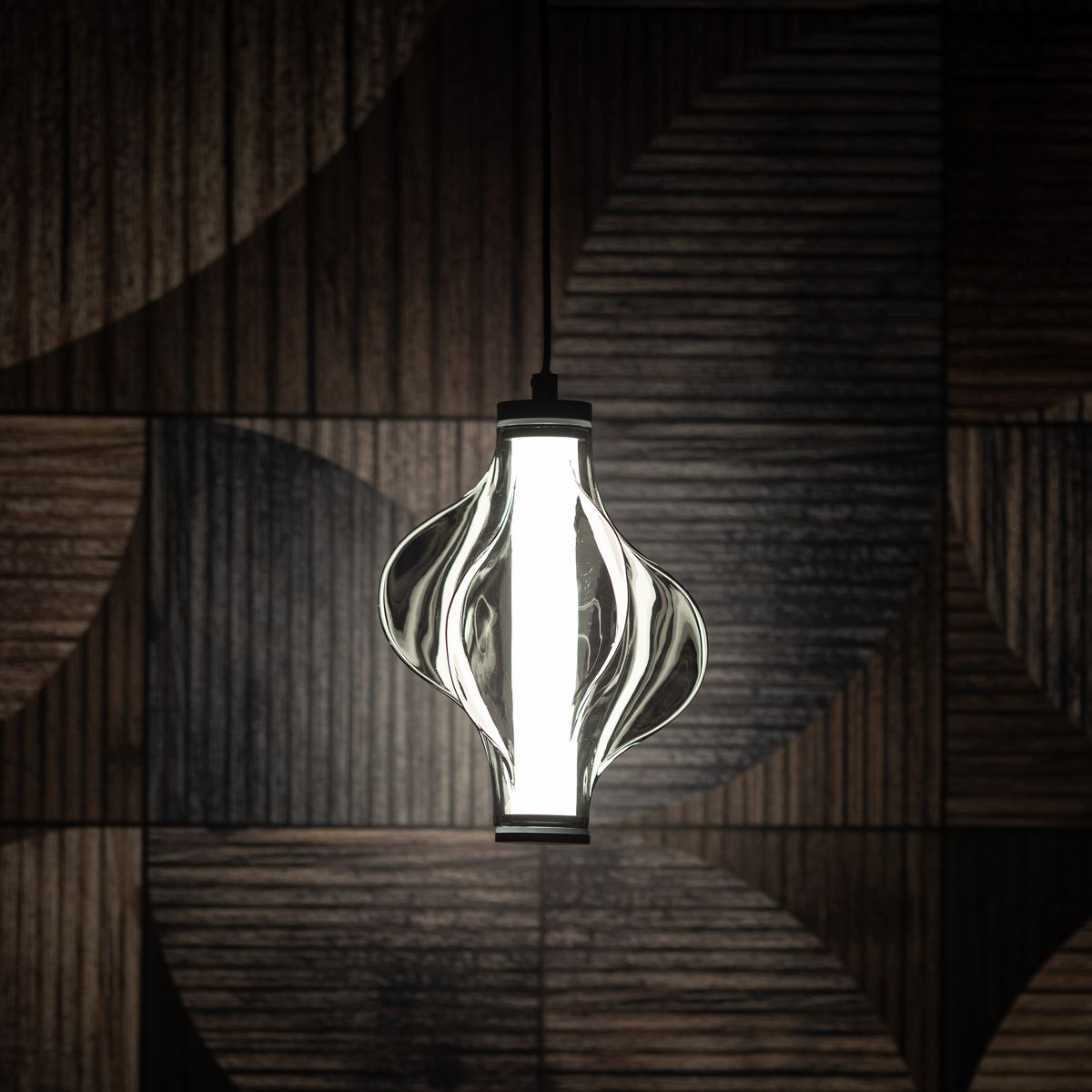 Lampa wisząca LED Elva o mocy 8W, kolor czarny/grafitowy, z unikalnym, falistym kloszem szklanym. Zawieszona na czarnym przewodzie, emituje ciepłe światło 3000K. Tło w odcieniach drewna podkreśla nowoczesny design.
