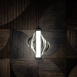 Lampa wisząca LED Elva o mocy 8W, kolor czarny/grafitowy, z unikalnym, falistym kloszem szklanym. Zawieszona na czarnym przewodzie, emituje ciepłe światło 3000K. Tło w odcieniach drewna podkreśla nowoczesny design.