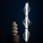 Lampa wisząca LED Elva 22W 3000K w kolorze czarnym i grafitowym, o eleganckim kształcie z przezroczystymi elementami, zawieszona w pionie. W tle widoczny ceramiczny wazon w odcieniu beżu na drewnianej podstawie. Tło ciemnoniebieskie.