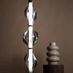 Lampa wisząca LED Elva 22W 3000K o czarnej/grafitowej obudowie, z efektowną, falistą konstrukcją. Zbliżenie na lampę w pionowej pozycji, z widocznym ciepłym światłem. Tło w neutralnym, brązowym odcieniu, obok ceramiczna dekoracja.