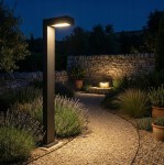 Czarna lampa stojąca Hebin H60 z gniazdem GX53, umieszczona na ścieżce ogrodowej w otoczeniu roślinności. Lampa emituje ciepłe światło, oświetlając kamienny mur w tle, na tle nocnego nieba.