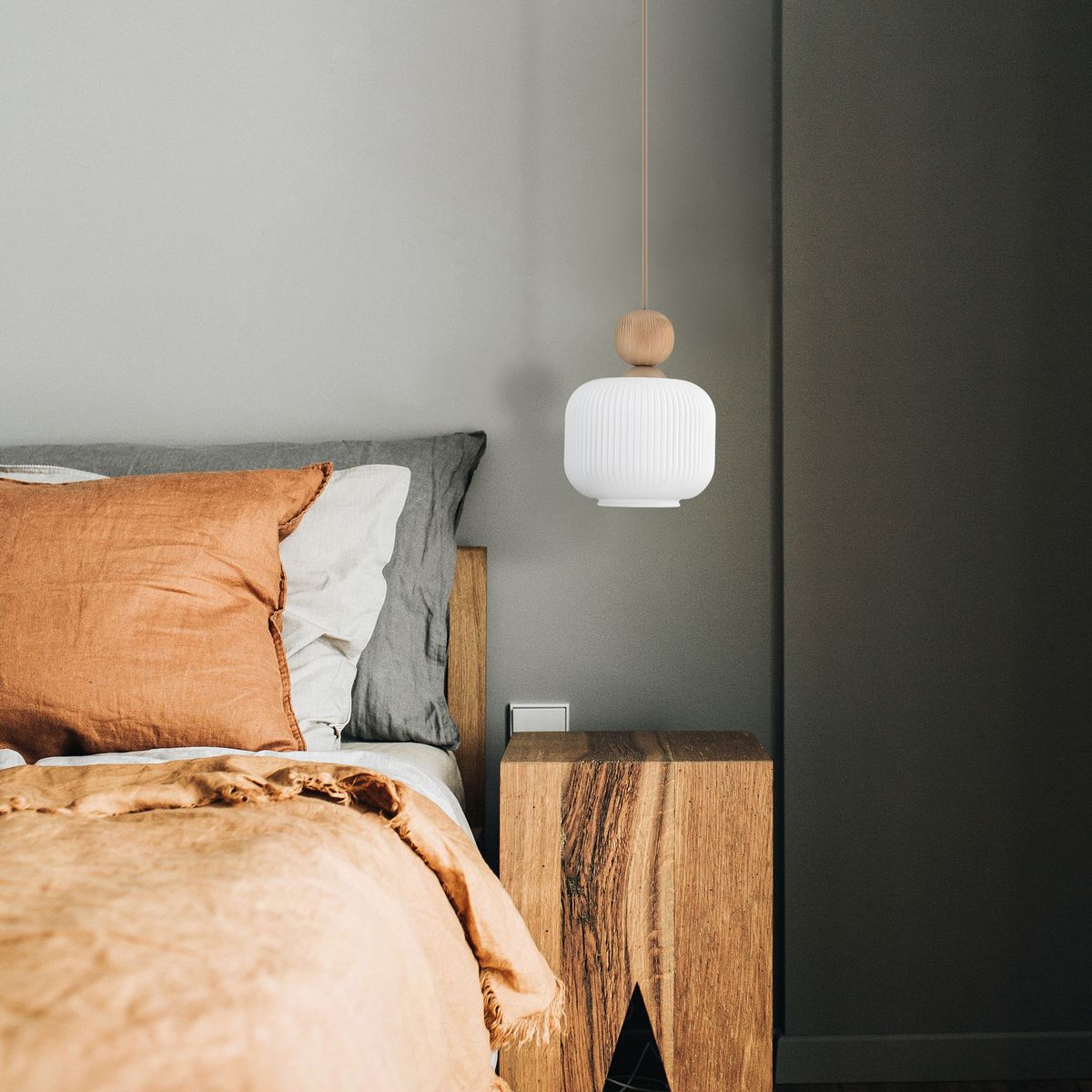 Lampa wisząca Ballo w kolorze beżowym z kloszem w kształcie cylindrycznym, z drewna, zawieszona na cienkim przewodzie. Widoczna w aranżacji sypialni nad łóżkiem z brązowymi poduszkami i drewnianą szafką nocną.