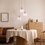 Lampa wisząca Ballo w kolorze beżowym z trzema kloszami w kształcie sześcianu, wykonana z matowego szkła, zawieszona na cienkich linkach. Elementy drewniane u góry kloszy. Produkt w aranżacji z okrągłym stołem, krzesłem i dekoracjami w tle.
