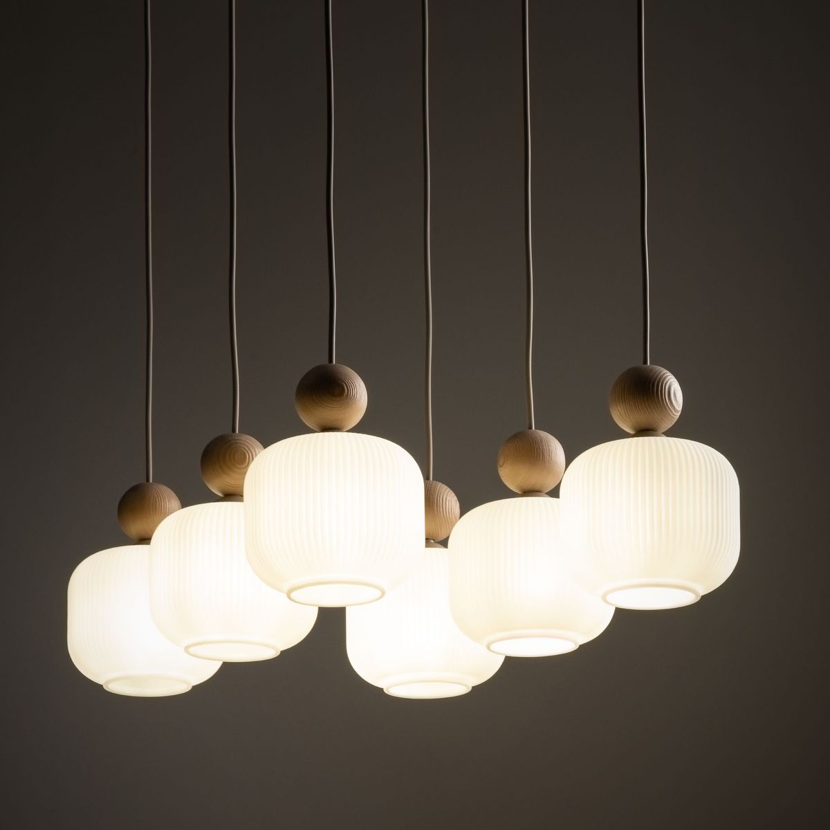 Ballo lampa wisząca z sześcioma kloszami E14 w kolorze beżowym. Klosze mają delikatne rowki, a u góry znajdują się drewniane kulki. Lampa zawieszona na cienkich, białych przewodach, prezentuje się nowocześnie na ciemnym tle.