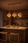 Beżowa lampa wisząca Sisu z trzema oprawami E27, zawieszona nad stołem w kuchni. Metalowy stelaż z kloszami w kolorze beżowym. W tle widoczne drewniane meble oraz naczynia, w tym miska z zielonymi jabłkami.