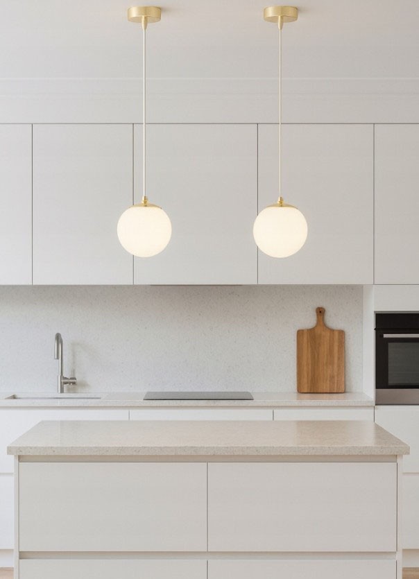 Lampa wisząca Sphere w kolorze złotym z białym, okrągłym kloszem, zawieszona na długim przewodzie. Widok z góry na nowoczesną kuchnię z białymi meblami i drewnianą deska do krojenia w tle. Dwie lampy umieszczone równolegle nad wyspą kuchenną.