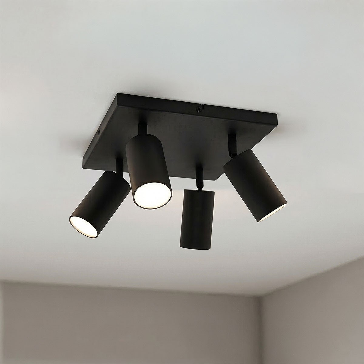 Czarna lampa sufitowa Spoti SQ z czterema ruchomymi reflektorami na kwadratowej podstawie. Reflektory mają cylindryczny kształt, emitują ciepłe światło, zamontowane na suficie w pomieszczeniu. Tło w jasnym kolorze.