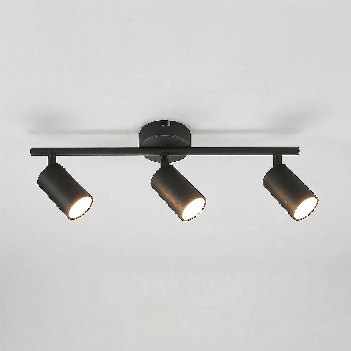 Czarna lampa sufitowa typu spot z trzema ruchomymi reflektorami na metalowej ramie. Reflektory mają cylindryczny kształt, emitują ciepłe światło. Umożliwia kierunkowe oświetlenie, zamontowana na okrągłej podstawie. Idealna do nowoczesnych wnętrz.