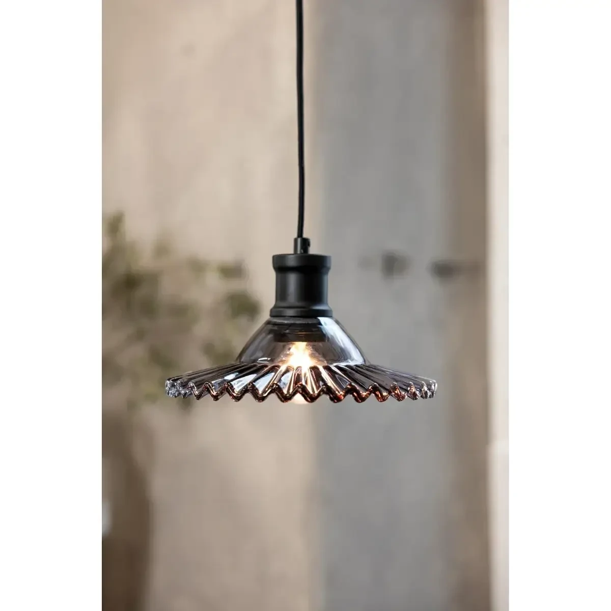 Lampa wisząca Novia E27 w kolorze czarnym z dymionym kloszem. Klosz o falistym kształcie, przezroczysty, wykonany ze szkła. Produkt zawieszony na czarnym przewodzie, w tle neutralne otoczenie.