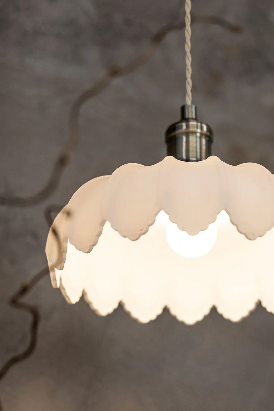 Lampa wisząca Kurage E27 w kolorze antycznego mosiądzu z białym kloszem o falistym kształcie. Widoczne ciepłe światło żarówki. Produkt zawieszony na plecionym przewodzie, prezentowany z bliskiej odległości. Tło w stonowanej kolorystyce.
