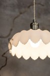 Lampa wisząca Kurage E27 w kolorze antycznego mosiądzu z białym kloszem o falistym kształcie. Widoczne ciepłe światło żarówki. Produkt zawieszony na plecionym przewodzie, prezentowany z bliskiej odległości. Tło w stonowanej kolorystyce.