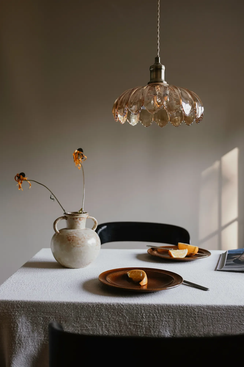 Lampa wisząca Kurage E27 w antycznym mosiądzu z bursztynowym kloszem w kształcie kwiatów, zawieszona nad stołem. W tle widoczny ceramiczny wazon i talerze z cytrusami na drewnianych talerzach. Proste, stonowane tło.