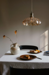 Lampa wisząca Kurage E27 w antycznym mosiądzu z bursztynowym kloszem w kształcie kwiatów, zawieszona nad stołem. W tle widoczny ceramiczny wazon i talerze z cytrusami na drewnianych talerzach. Proste, stonowane tło.