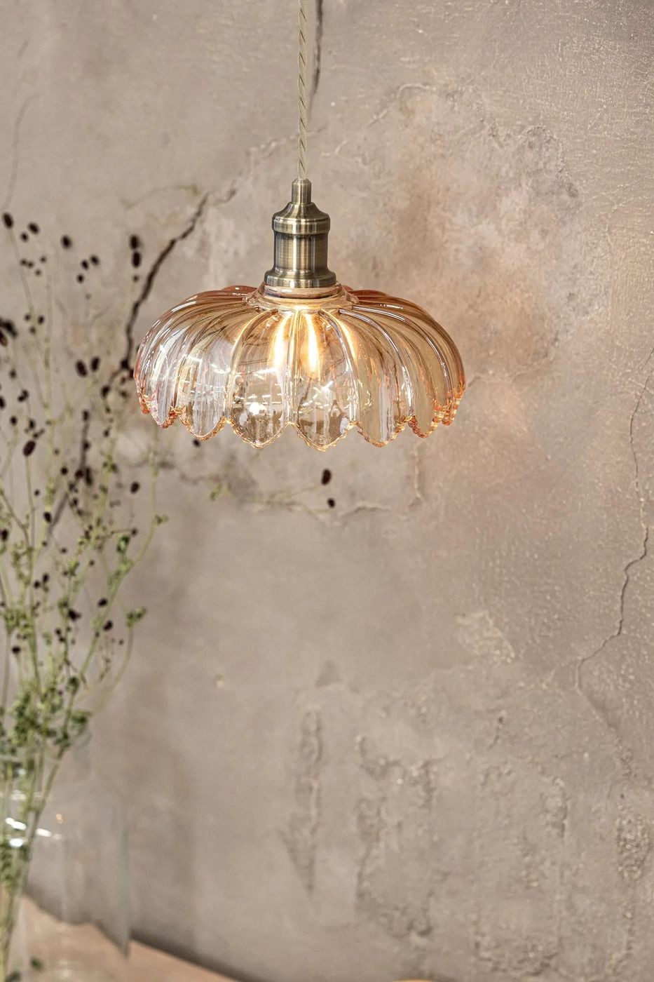 Lampa wisząca Kurage w kolorze antycznego mosiądzu z bursztynowym kloszem o falistej formie. Klosz wykonany ze szkła, zawieszony na cienkim przewodzie. Tło w odcieniach szarości, z delikatnymi roślinami w tle.