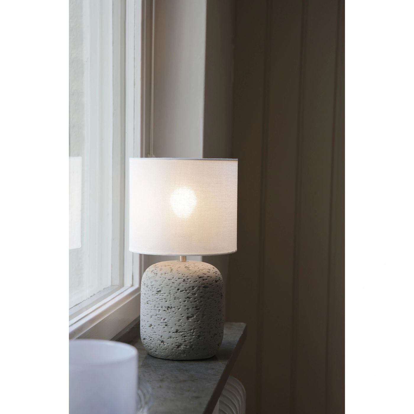 Lampa stołowa Montagna E14 w odcieniu szarym z białym abażurem. Posiada okrągłą, teksturowaną podstawę i proste, eleganckie wykończenie. Ustawiona na parapecie, emituje ciepłe światło, podkreślając minimalistyczny styl wnętrza.