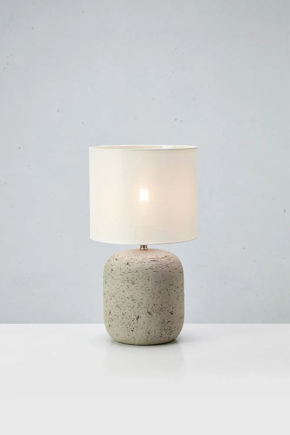 Lampa stołowa Montagna E27 w odcieniu szaro-białym z okrągłą, betonową podstawą i białym, materiałowym kloszem. Produkt stoi na białym blacie, w prostym, neutralnym tle, co podkreśla jego minimalistyczny styl.