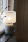 Lampa stołowa Montagna E27 w szarej ceramice z okrągłym, białym abażurem. Ujęcie z boku, stojąca na parapecie. Widoczna żarówka wewnątrz abażuru. Prosta, minimalistyczna forma, idealna do nowoczesnych wnętrz.