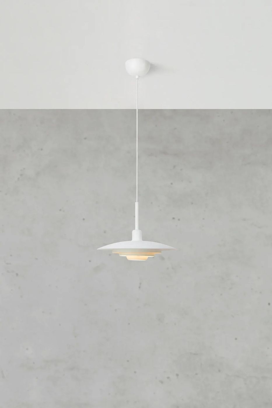 Lampa wisząca E27 w kolorze białym, z okrągłym, wielowarstwowym kloszem. Zawieszona na cienkim przewodzie, umieszczona na tle szarej, betonowej ściany. Klosz ma lekko zaokrąglony kształt, co podkreśla nowoczesny design.