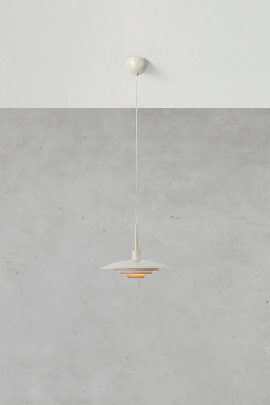 Beżowa lampa wisząca E27 z okrągłym kloszem, zawieszona na cienkim przewodzie. Klosz składa się z trzech warstw, a wewnętrzna część ma ciepły, pomarańczowy odcień, co tworzy przytulny efekt świetlny. Lampa montowana do sufitu na prostym mocowaniu.