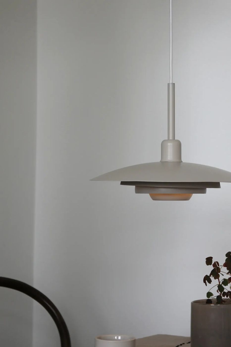 Piny lampa wisząca E27 w kolorze beżowym, z okrągłym kloszem o dwóch warstwach, zawieszona na białym przewodzie. Widoczna w minimalistycznym wnętrzu, obok filiżanki i doniczki z rośliną. Kąt ujęcia z boku, ukazujący detal oraz nowoczesny design.