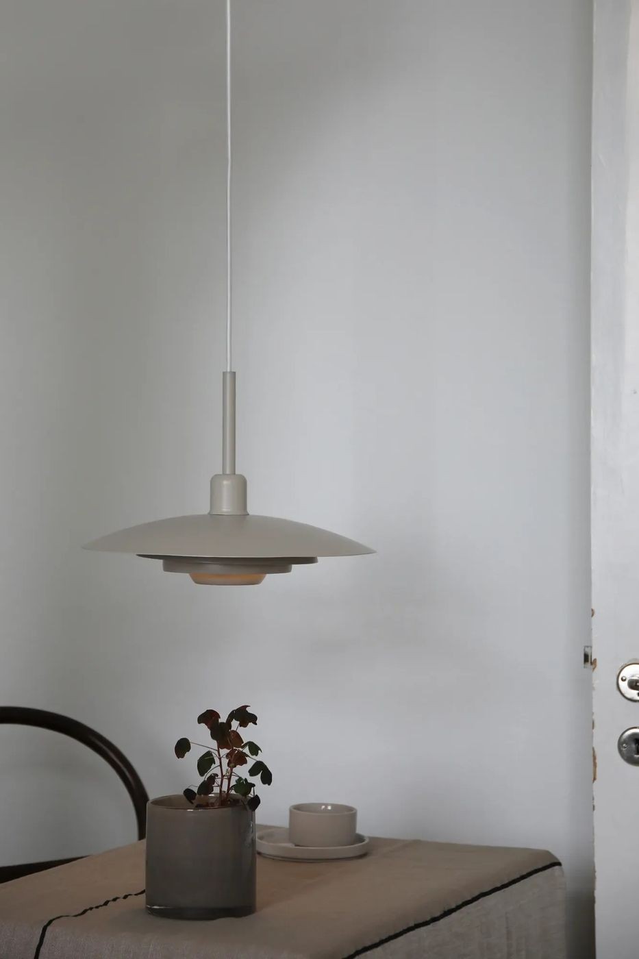 Beżowa lampa wisząca E27 Piny z metalowym kloszem o szerokim, płaskim kształcie, zawieszona na cienkim przewodzie. W tle stół z szarym doniczką i filiżanką na jasnym obrzeżu, na gładkiej, jasnoszarej ścianie.