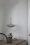 Beżowa lampa wisząca E27 Piny z metalowym kloszem o szerokim, płaskim kształcie, zawieszona na cienkim przewodzie. W tle stół z szarym doniczką i filiżanką na jasnym obrzeżu, na gładkiej, jasnoszarej ścianie.