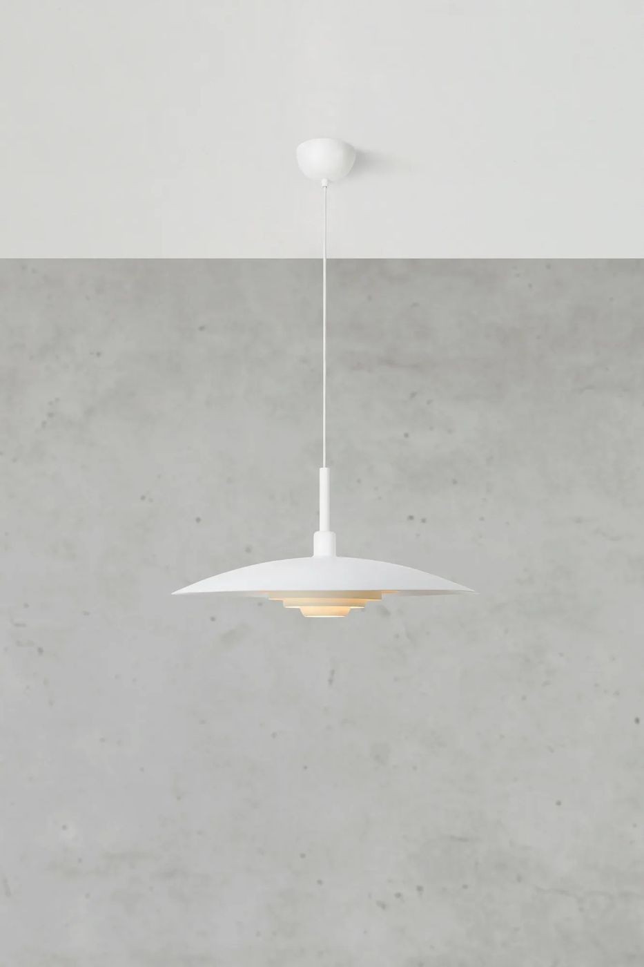 Lampa wisząca E27 w kolorze białym, z okrągłym kloszem o warstwowej konstrukcji. Zawieszona na cienkim przewodzie, umieszczona pod sufitem w jasnym otoczeniu betonu, z delikatnym podświetleniem wewnętrznym.
