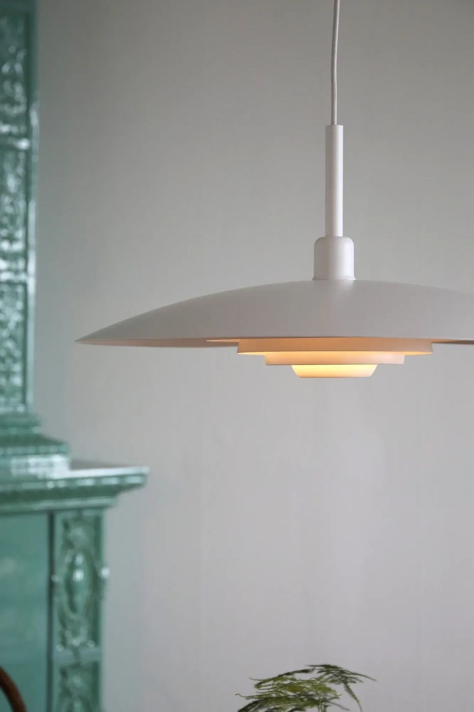 Lampa wisząca E27 Piny w kolorze białym, o okrągłym kloszu w kształcie talerza, zawieszona na cienkim przewodzie. Widoczny ciepły blask emitowany przez wewnętrzny element. Tło w stonowanej kolorystyce, z elementami dekoracyjnymi w tle.