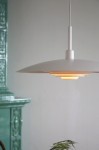 Lampa wisząca E27 Piny w kolorze białym, o okrągłym kloszu w kształcie talerza, zawieszona na cienkim przewodzie. Widoczny ciepły blask emitowany przez wewnętrzny element. Tło w stonowanej kolorystyce, z elementami dekoracyjnymi w tle.