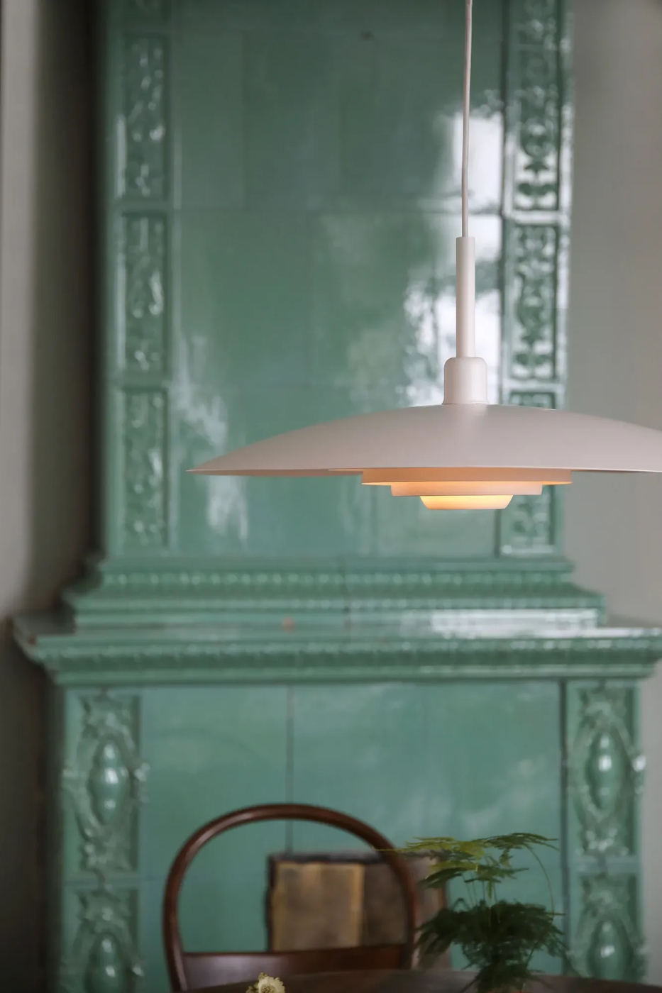 Lampa wisząca E27 w kolorze białym, o okrągłej, płaskiej formie z subtelnym, złotym wykończeniem wewnątrz. Zawieszona na cienkim, białym kablu, umieszczona nad drewnianym stołem w aranżacji z zieloną ścianą w tle.