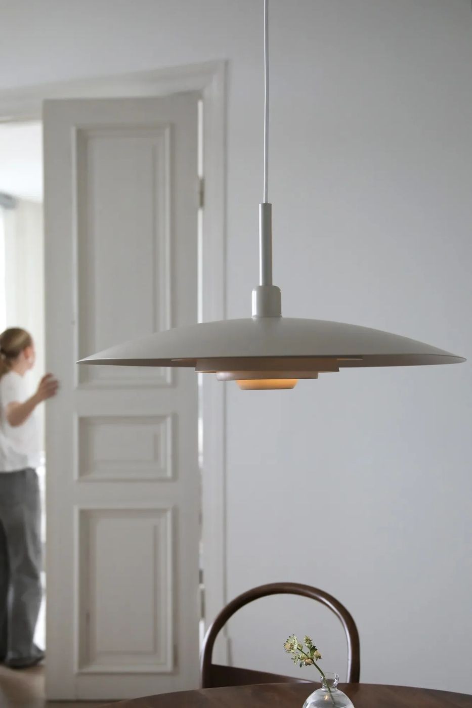 Beżowa lampa wisząca E27 z metalowym kloszem w kształcie talerza, zawieszona na białym przewodzie. Ujęcie z boku, w tle widoczne otwarte drzwi i stół z wazonem. Klosz lekko osłania źródło światła, tworząc subtelne oświetlenie.