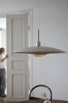 Beżowa lampa wisząca E27 z metalowym kloszem w kształcie talerza, zawieszona na białym przewodzie. Ujęcie z boku, w tle widoczne otwarte drzwi i stół z wazonem. Klosz lekko osłania źródło światła, tworząc subtelne oświetlenie.