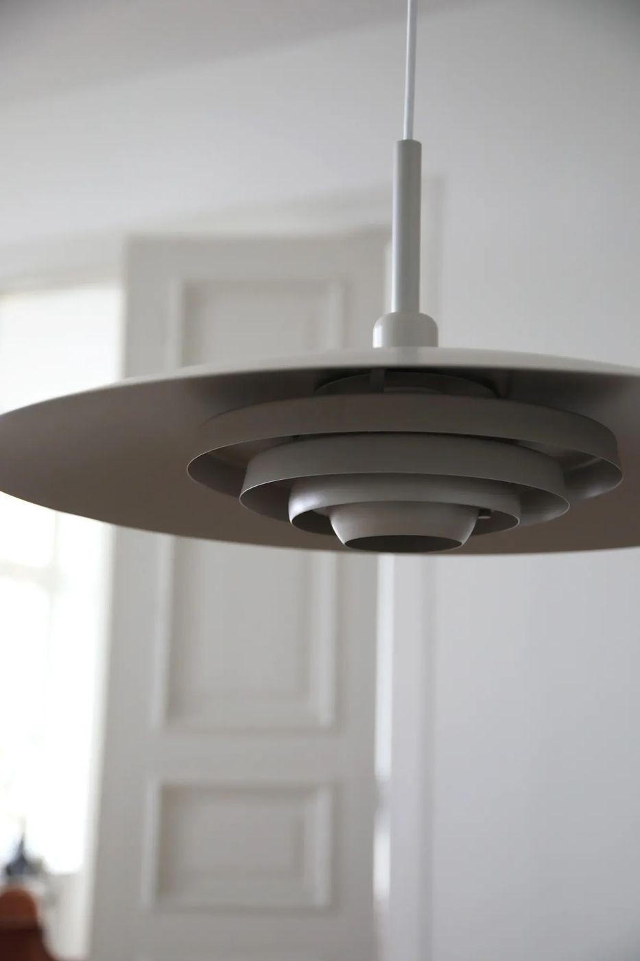Beżowa lampa wisząca E27 z okrągłym, płaskim kloszem i warstwową konstrukcją, zawieszona na białym przewodzie. Widok z dołu, ukazujący detale wewnętrzne klosza. Tło w jasnym wnętrzu z drzwiami w stylu klasycznym.