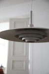 Beżowa lampa wisząca E27 z okrągłym, płaskim kloszem i warstwową konstrukcją, zawieszona na białym przewodzie. Widok z dołu, ukazujący detale wewnętrzne klosza. Tło w jasnym wnętrzu z drzwiami w stylu klasycznym.