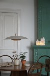 Beżowa lampa wisząca E27 w kształcie talerza, zawieszona na cienkim, białym kablu. Widoczna w aranżacji na okrągłym stole, otoczona roślinami i ceramicznymi akcesoriami. Tło jasne z drewnianymi elementami i zielonym kominkiem.