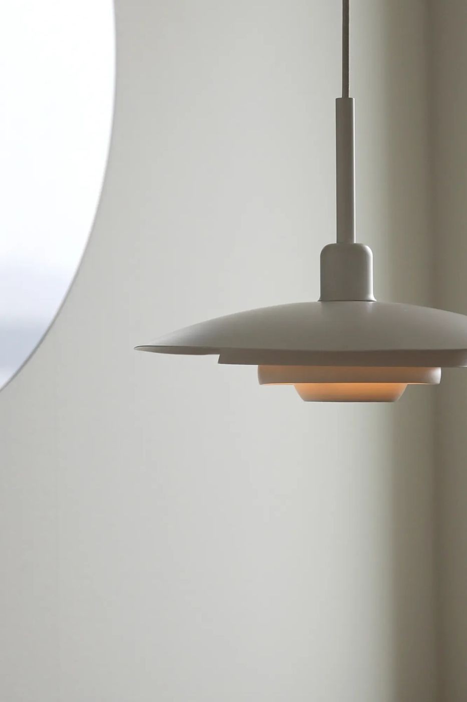 Lampa wisząca E27 w kolorze białym, o nowoczesnym kształcie. Zawieszona na cienkim kablu, z okrągłym kloszem, który ma wycięcia. Tło jasne, gładkie, co podkreśla minimalistyczny design lampy.