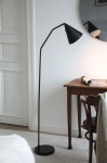 Czarna lampa podłogowa LED Funil 4,9W, z cienką, giętką nogą i stożkowym kloszem, ustawiona obok drewnianego stolika. Lampa ma okrągłą podstawę i jest umieszczona na jasnym dywaniku w minimalistycznym wnętrzu.