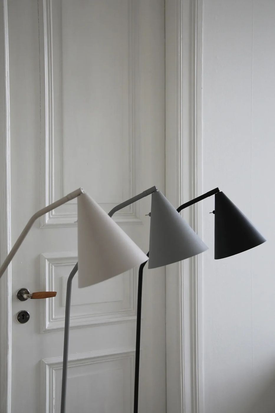 Lampa podłogowa LED Funil 4,9W w kolorze beżowym, z prostym, metalowym stelażem. Klosz w kształcie stożka, skierowany w dół, umieszczony na łuku. Obok klosze w kolorach szarym i czarnym. Tło jasne, minimalistyczne, podkreślające elegancję lamp.