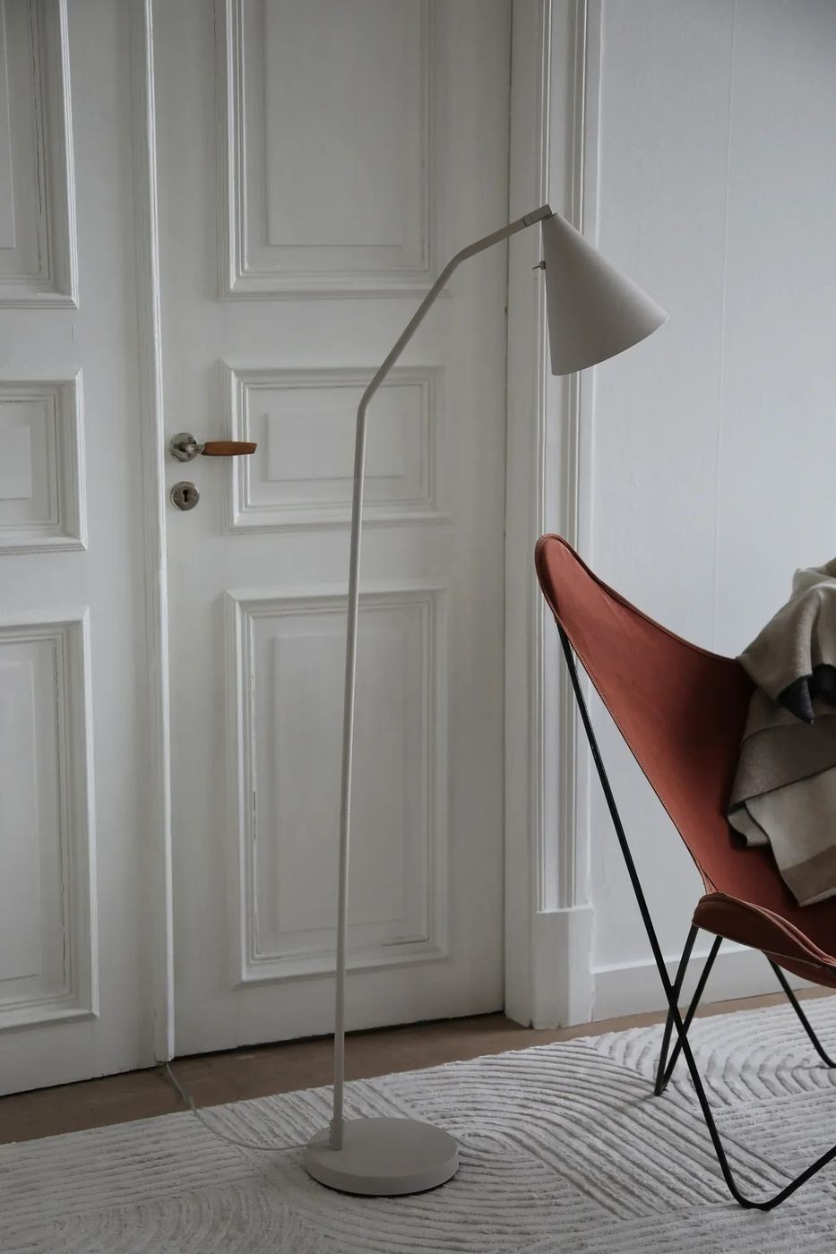 Lampa podłogowa LED Funil 4,9W w beżowej kolorystyce, o smukłej, zakrzywionej nodze i stożkowym kloszu. Ustawiona obok czerwonego fotela na tle białych drzwi. Dodatkowo widoczna okrągła podstawa i kabel zasilający.