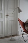 Lampa podłogowa LED Funil 4,9W w beżowej kolorystyce, o smukłej, zakrzywionej nodze i stożkowym kloszu. Ustawiona obok czerwonego fotela na tle białych drzwi. Dodatkowo widoczna okrągła podstawa i kabel zasilający.