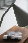 Lampa podłogowa LED Funil 4,9W w kolorze szarym, z metalowym kloszem w kształcie stożka. Widok z boku, zbliżenie na głowicę lampy i elementy mocujące. Na drewnianym stoliku widoczny magazyn i fragment tuby.