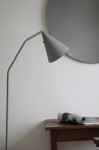 Lampa podłogowa LED Funil 4,9W w kolorze szarym z metalowym kloszem w kształcie stożka, zamontowana na cienkiej, metalowej nodze. W tle widoczna okrągła lustro na jasnej ścianie oraz drewniany stół z przewróconym czasopismem.