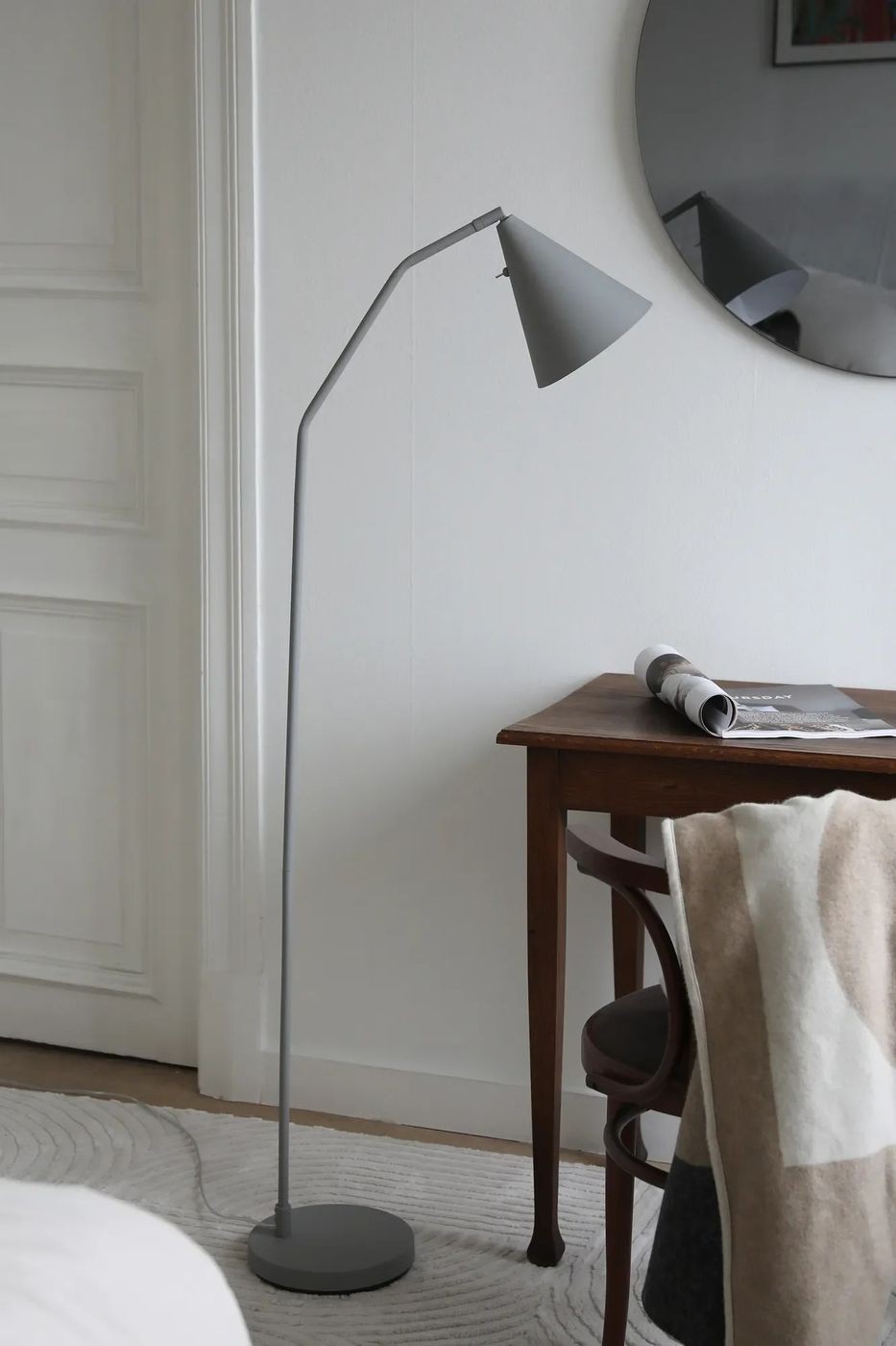 Lampa podłogowa LED Funil 4,9W w kolorze szarym, z prostą, metalową nogą i stożkowym kloszem. Ustawiona obok drewnianego stołu, na tle gładkiej, jasnej ściany oraz okrągłego lustra. Widoczny także kawałek dywanu i złożony koc.