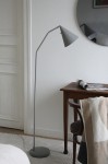Lampa podłogowa LED Funil 4,9W w kolorze szarym, z prostą, metalową nogą i stożkowym kloszem. Ustawiona obok drewnianego stołu, na tle gładkiej, jasnej ściany oraz okrągłego lustra. Widoczny także kawałek dywanu i złożony koc.