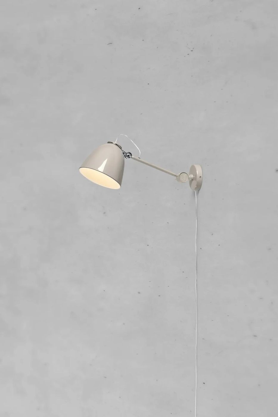 Kinkiet Tilt E27 w kolorze szarym, z metalowym kloszem w kształcie stożka, zamocowany na ścianie. Lampa ma regulowany wysięgnik oraz widoczny kabel z włącznikiem na ścianie. Tło w odcieniach szarości.
