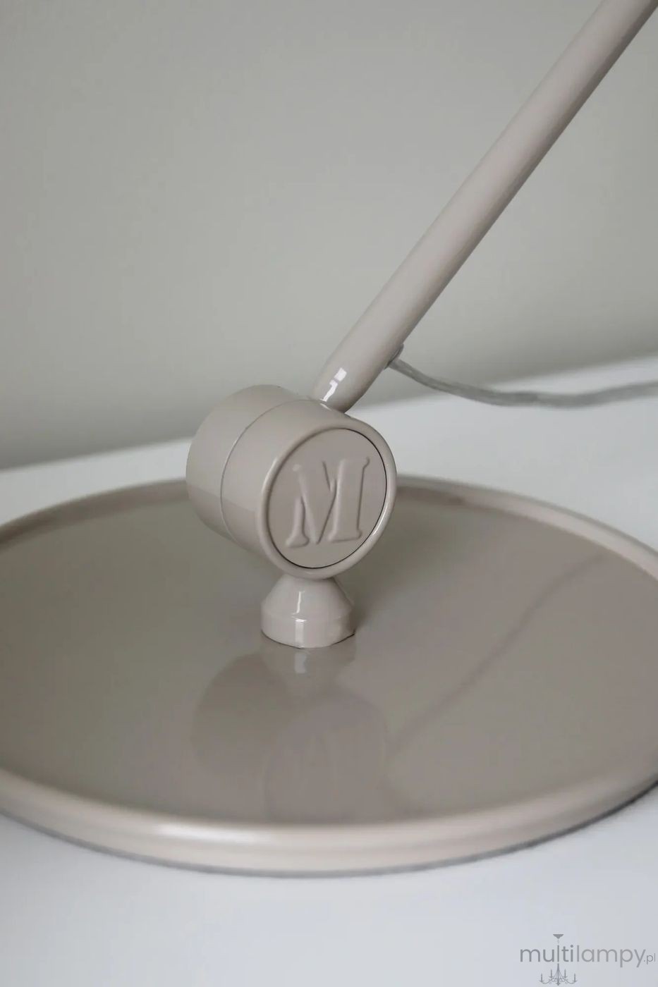 Lampa stołowa Tilt E27 w kolorze szarym z okrągłą podstawą. Ujęcie z boku ukazuje regulowany ramion z ozdobnym logo M. Gładka, matowa powierzchnia, kablowe zasilanie. Idealna do biura lub domowego wnętrza.