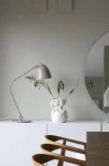 Szara lampa stołowa Tilt E27 z metalowym kloszem, z regulowanym ramieniem, ustawiona na białym meblu obok białego wazonu z roślinami. W tle widoczna okrągła lustro na szarej ścianie.