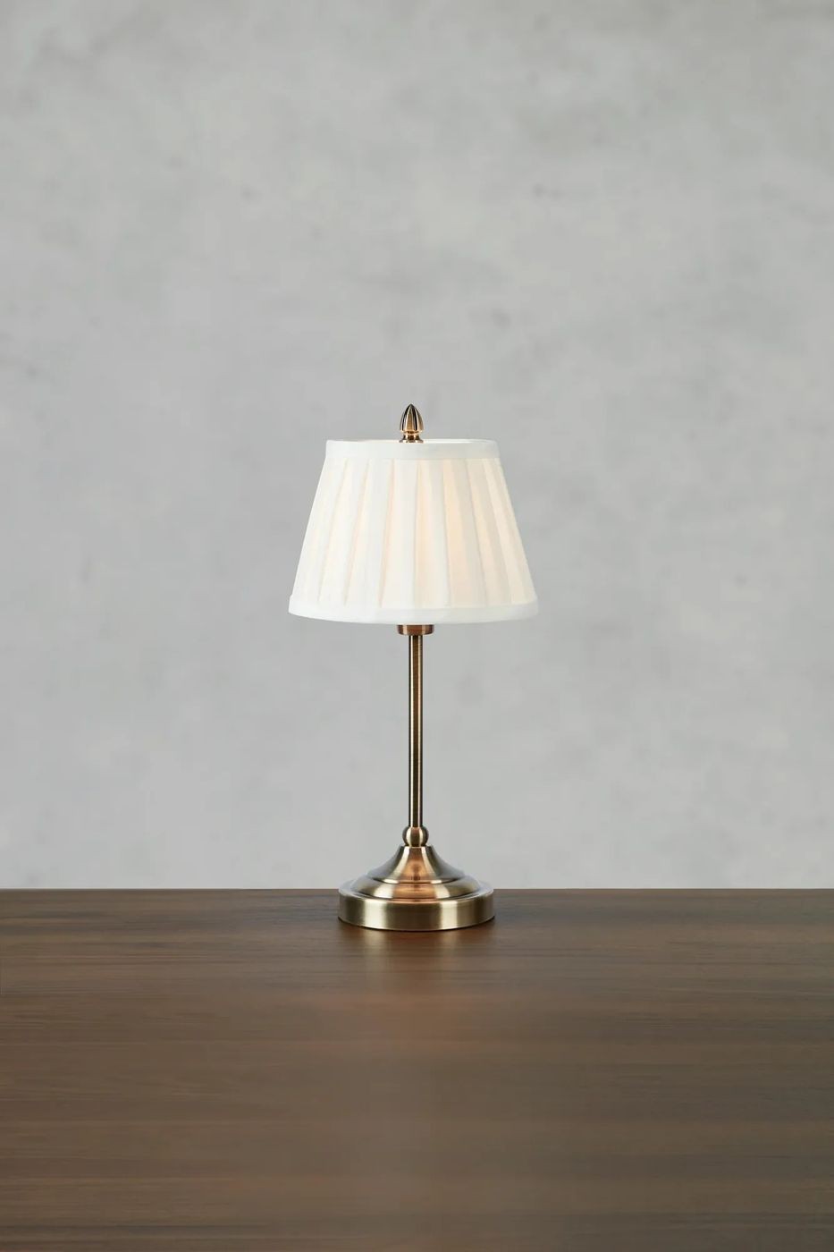 Lampa stołowa Oxford E14 w antycznym mosiądzu z białym, plisowanym kloszem. Ustawiona na drewnianym blacie, w frontalnym ujęciu. Klosz ma kształt stożka, a podstawa lampa jest okrągła i błyszcząca. Tło w odcieniach szarości.